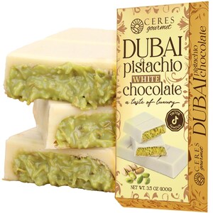 May include: White chocolate bars with a pistachio filling are stacked next to a box of Dubai Pistachio White Chocolate. The box has gold and brown accents and the text "Dubai Pistachio White Chocolate" and "a taste of luxury". The net weight is 3.5 oz (100g).