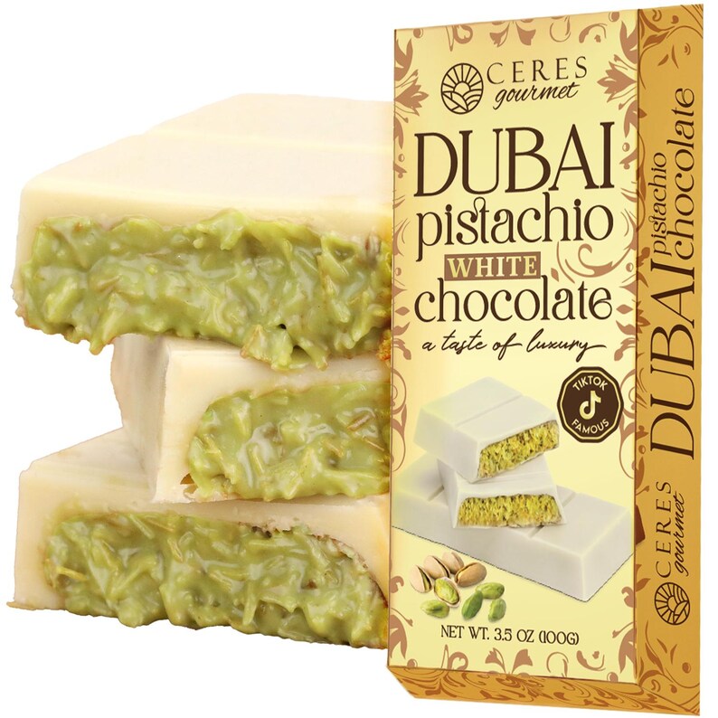 May include: White chocolate bars with a pistachio filling are stacked next to a box of Dubai Pistachio White Chocolate. The box has gold and brown accents and the text "Dubai Pistachio White Chocolate" and "a taste of luxury". The net weight is 3.5 oz (100g).