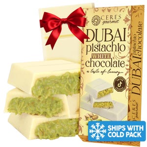 May include: White chocolate bars with a pistachio filling are stacked in front of a box of Dubai Pistachio White Chocolate. A red bow adorns a white gift box. The packaging includes the text "Dubai Pistachio White Chocolate" and "a taste of luxury."