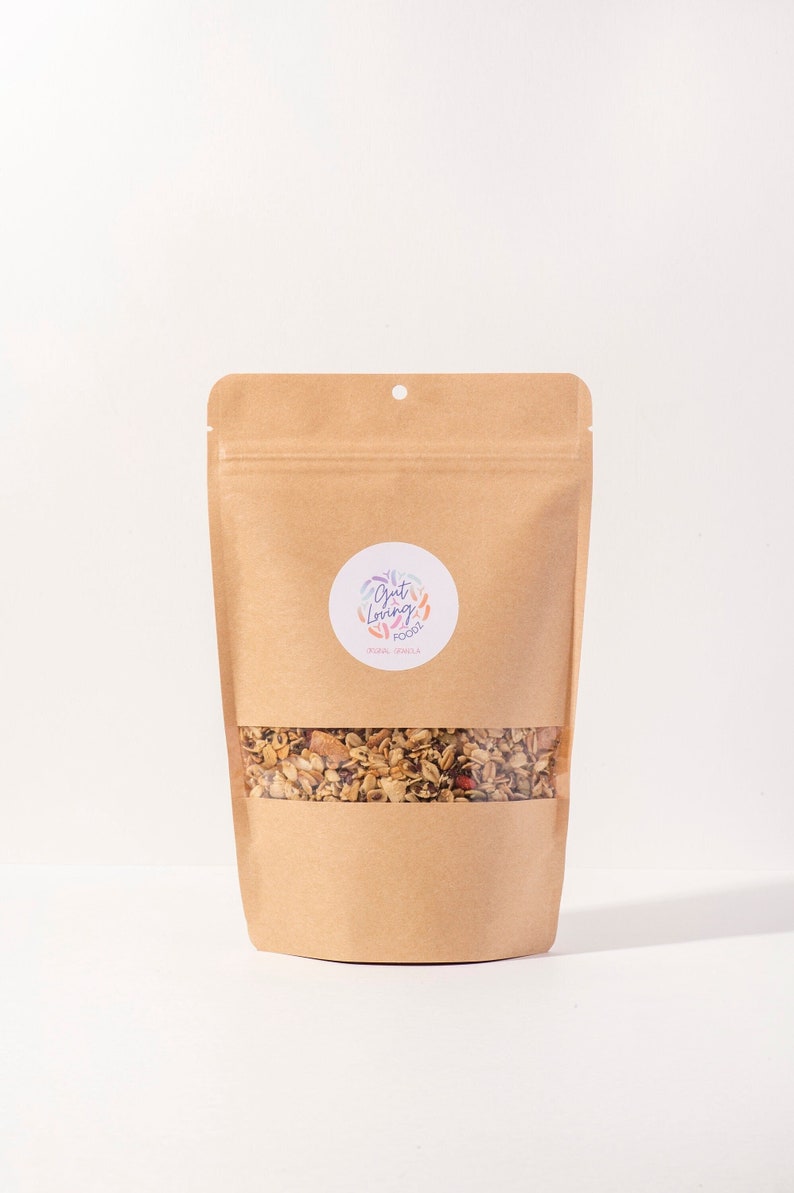 May include: A brown paper bag of granola with a clear window, showing the contents. The granola contains various grains, nuts, and dried fruits. A round sticker with the text "Gut Loving Foods" is on the front.