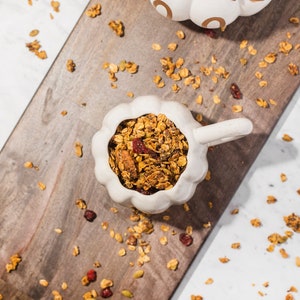 May include: A white ceramic pumpkin-shaped bowl filled with granola, set on a wooden chopping board. The granola includes oats, nuts, seeds, and dried cranberries. Another pumpkin-shaped ceramic piece is visible.