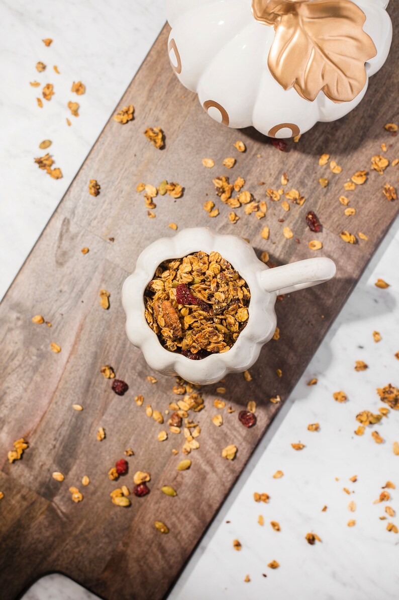 May include: A white ceramic pumpkin-shaped bowl filled with granola, set on a wooden chopping board. The granola includes oats, nuts, seeds, and dried cranberries. Another pumpkin-shaped ceramic piece is visible.