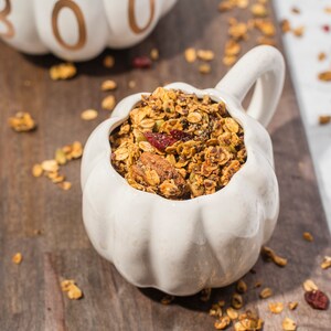 May include: A white pumpkin-shaped bowl filled with granola, containing oats, nuts, and dried cranberries. The bowl has a handle and is on a wooden surface. Another pumpkin-shaped object with the word "BOO" is in the background.