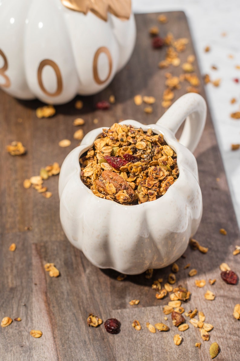 May include: A white pumpkin-shaped bowl filled with granola, containing oats, nuts, and dried cranberries. The bowl has a handle and is on a wooden surface. Another pumpkin-shaped object with the word "BOO" is in the background.