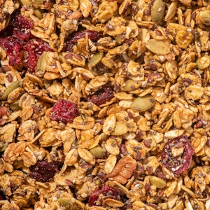 May include: Close-up of a mix of granola with oats, dried cranberries, pumpkin seeds, chia seeds, and pecans. The granola has a golden-brown colour with red and green accents, creating a textured and appetising appearance.