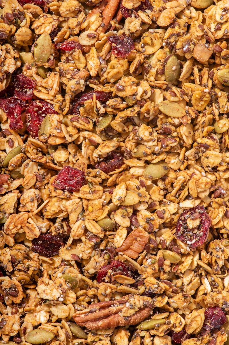 May include: Close-up of a mix of granola with oats, dried cranberries, pumpkin seeds, chia seeds, and pecans. The granola has a golden-brown colour with red and green accents, creating a textured and appetising appearance.