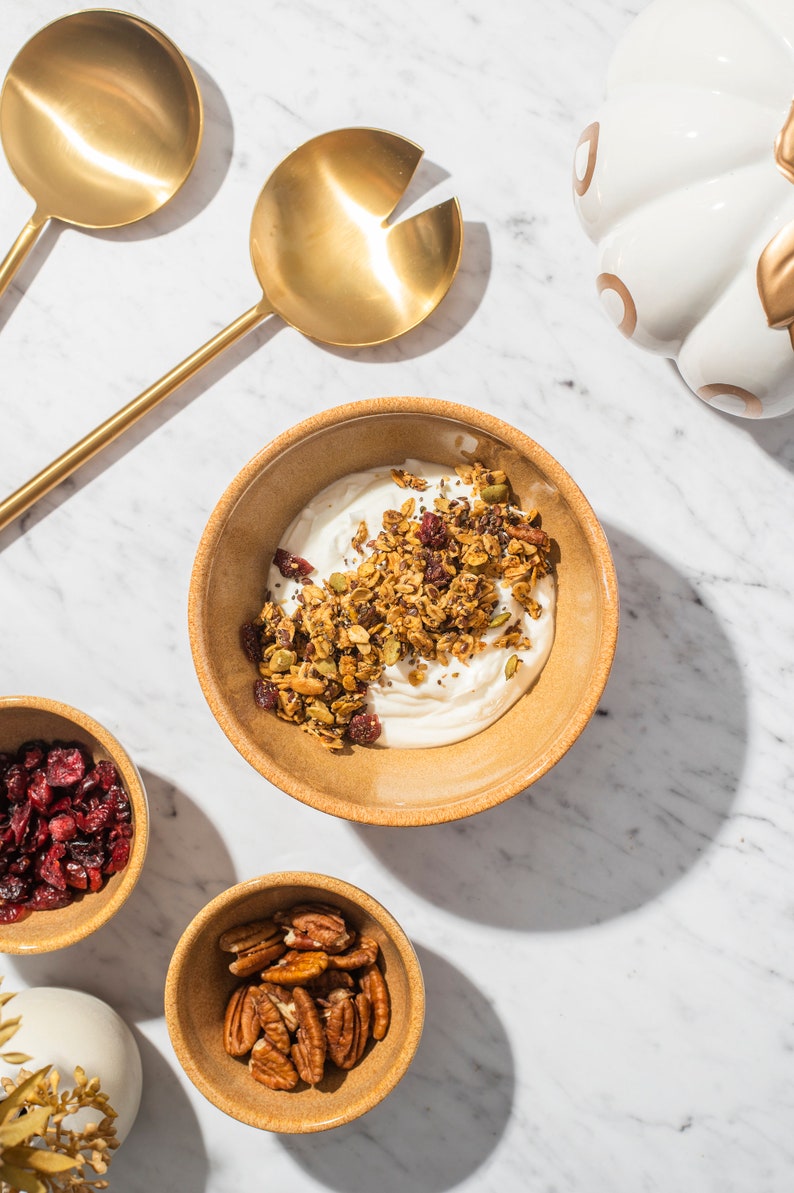May include: A breakfast scene featuring a bowl of yogurt topped with granola, dried cranberries, and pecans. Two gold serving spoons and a decorative pumpkin are also present. The items are on a white marble surface.