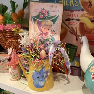 May include: A yellow ceramic watering can with a blue bunny design, surrounded by colorful Easter grass and a vintage Easter card that says "Happy Easter".
