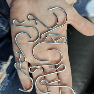 May include: A collection of silver-colored metal S-hooks and clips. The hooks have a curved design, and the clips have a straight bar with a small loop. The hooks and clips are arranged in a hand.