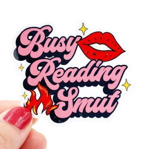 May include: Pink and black sticker with the text "Busy Reading Smut" in a retro font. A red cartoon mouth with white teeth is next to the text. There are small yellow stars around the text.