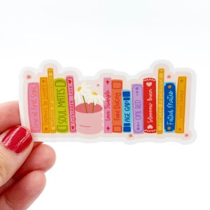 May include: A sticker of a bookshelf with a variety of colorful books. The books have titles such as "Love at First Sight", "Soul Mates", "Forbidden Love", "Fake Dating", "Age Gap", "One Bed", "Sloooow Burn", "Forced Proximity", "Fated Mates", and "Second Chance". There is a small white flower pot with a white daisy in it on the shelf.