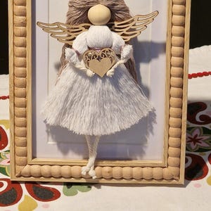 Puede incluir: Una figurita de ángel de hilo blanco que sostiene un corazón de madera con un diseño rosa y dorado. El ángel está enmarcado en un marco de madera claro con un borde de cuentas.