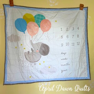 May include: A white and blue baby quilt with a gray elephant holding balloons. The balloons are pink, blue, green, and yellow. The quilt has a number chart for days, weeks, months, and years. The text "April Dawn Quilts" is at the bottom of the quilt.