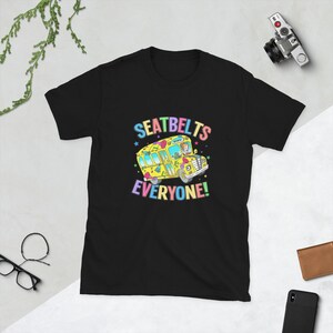 Op de afbeelding: Een zwart t-shirt met korte mouwen en een afbeelding van een gele schoolbus met een bestuurder en passagier. De tekst SEATBELTS EVERYONE! staat in kleurrijke letters boven en onder de bus, omringd door kleine sterren en muzieknoten.