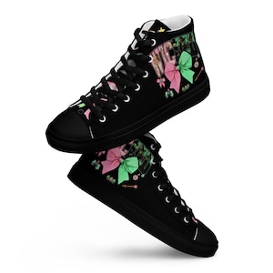Op de afbeelding: Zwarte hoge sneakers met witte veters. De sneakers hebben een ontwerp met roze en groene strikken en het woord "VICTORIA" in het groen. De schoenen hebben een zwarte zool en een witte binnenkant.