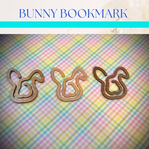 May include: Three wooden bunny bookmarks on a pink and green checkered background. The bunny shapes are cut out of wood and have a natural wood color.