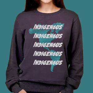 Puede incluir: Sudadera gris oscuro con la palabra "INDIGENOUS" repetida cinco veces en blanco, superpuesta a un mapa turquesa de América. La sudadera tiene mangas largas y cuello redondo.