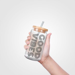 May include: A clear glass tumbler with a bamboo lid and a silver straw. The tumbler has the words "GOOD VIBES" printed in black. The tumbler is held in a hand against a white background.
