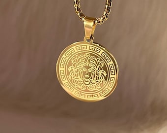 May include: A gold chain necklace with a round pendant featuring a detailed engraving of Medusa's head surrounded by a Greek key border.