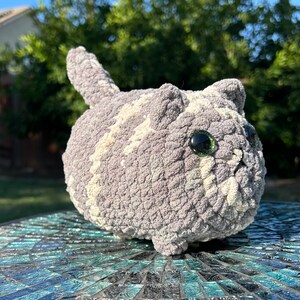Puede incluir: Un juguete de peluche de gato de crochet gris y blanco con grandes ojos negros. El juguete est&aacute; sentado sobre una superficie de mosaico azul y verde.