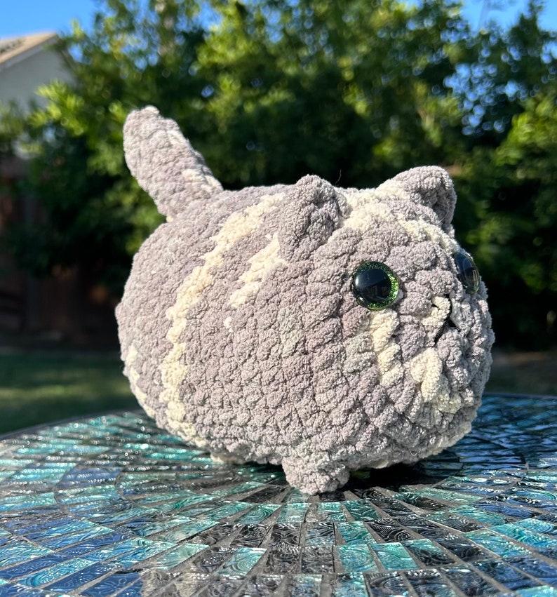 Puede incluir: Un juguete de peluche de gato de crochet gris y blanco con grandes ojos negros. El juguete est&aacute; sentado sobre una superficie de mosaico azul y verde.