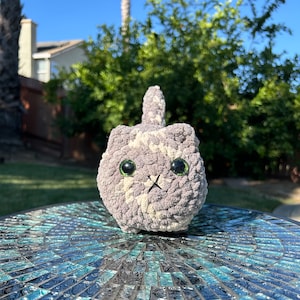 Puede incluir: Un juguete de peluche de gato de crochet gris y blanco con ojos verdes. El juguete est&aacute; sentado en una mesa de mosaico azul y negro.