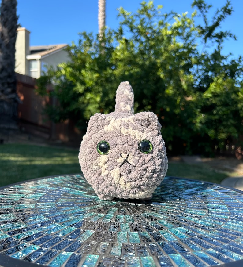 Puede incluir: Un juguete de peluche de gato de crochet gris y blanco con ojos verdes. El juguete est&aacute; sentado en una mesa de mosaico azul y negro.