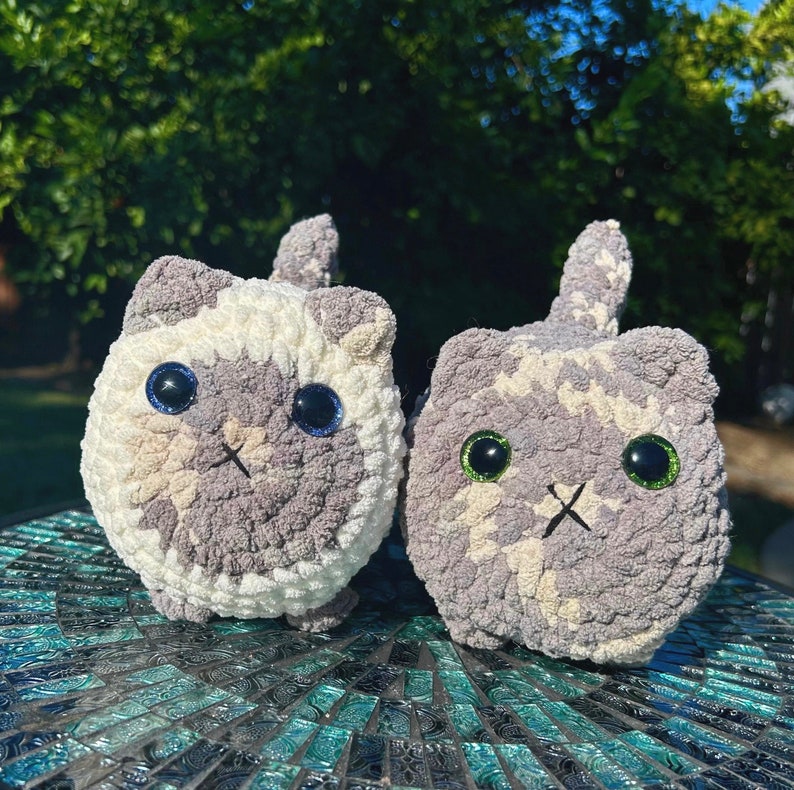 Puede incluir: Dos peluches de gato de crochet, uno gris y blanco con ojos azules, el otro gris con ojos verdes. Ambos est&aacute;n sentados sobre una superficie de mosaico.