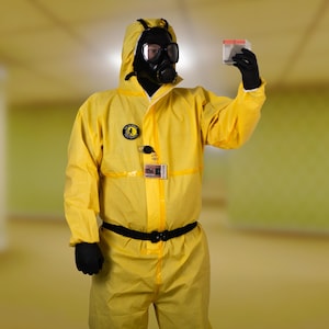 Peut inclure: Une personne porte une combinaison de protection jaune vif avec un masque à gaz et des gants noirs. La combinaison a une capuche et une ceinture noire. La personne tient une petite boîte blanche.