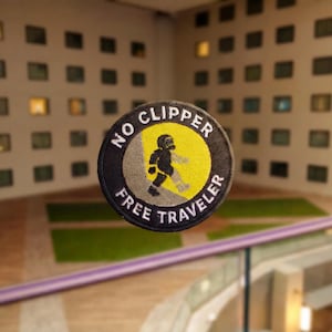 Może przedstawiać: Okrągła haftowana naszywka z czarną obwódką i białym napisem NO CLIPPER FREE TRAVELER. W środku znajduje się żółte koło z szarym ukośnym pasem, przedstawiające czarną sylwetkę osoby idącej pod górę pochyłości.