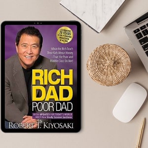 Puede incluir: Una tableta muestra la portada de "Rich Dad Poor Dad" de Robert T. Kiyosaki. La portada presenta una foto del autor y el t&iacute;tulo en amarillo. La tableta est&aacute; sobre un escritorio con un port&aacute;til, un rat&oacute;n y un posavasos tejido.