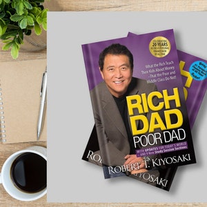 Puede incluir: Se muestran dos ejemplares de "Rich Dad Poor Dad" de Robert T. Kiyosaki. Las portadas de los libros presentan el retrato de un hombre sobre un fondo morado. El texto incluye "Celebrating 20 Years" y "#1 Personal Finance Book of All Time."