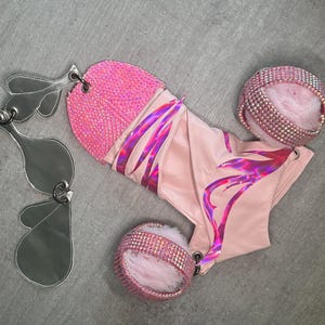 Peut inclure: Un vêtement rose clair avec une section supérieure recouverte de petits strass roses, des motifs de flammes irisées et deux poignets assortis ornés de strass roses et de fausse fourrure rose, accompagnés d'accessoires en métal argenté en forme d'ailes.