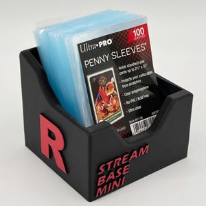 May include: A black card holder with a red "R" logo and the text "STREAM BASE MINI". It contains a stack of blue card sleeves labeled "Ultra PRO PENNY SLEEVES" that hold cards up to 2.5" x 3".