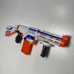 May include: A white and orange Nerf Retaliator Elite toy gun with a black grip and a blue and white logo. The gun has a magazine attached and a scope.