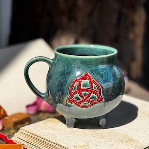 May include: A teal ceramic cauldron-shaped mug with a red Celtic knot design. The mug has a handle and three small feet. The mug is sitting on a stack of books with some rose petals and small brown objects.