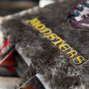 May include: A detailed shot of a book with a furry, dark gray cover. The word "MONSTERS" is embroidered in yellow on a black label. The book's spine and pages are visible, with red accents.