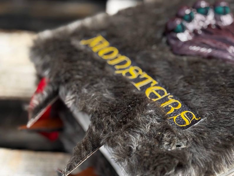 May include: A detailed shot of a book with a furry, dark gray cover. The word "MONSTERS" is embroidered in yellow on a black label. The book's spine and pages are visible, with red accents.