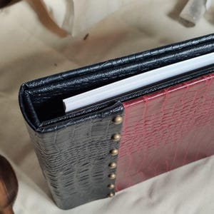 May include: A black and burgundy leather-bound journal with a textured cover and silver studs. The journal is closed and lying on a white surface.