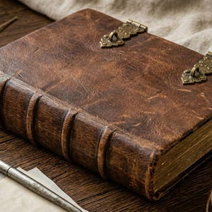 Könnte beinhalten: Ein dickes, rechteckiges Buch mit einem Einband aus strukturiertem, abgenutztem braunem Leder. Das Buch hat einen erhabenen Buchrücken mit vier horizontalen Bünden und zwei verzierte Messingschließen im Antik-Stil auf dem Vorderdeckel. Der Buchschnitt ist goldfarben.