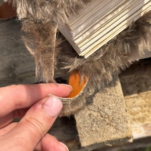 May include: Close-up of a section of brown faux fur with a stack of thin, light-colored wooden planks. The fur has a shaggy texture, and a small piece is being held up, revealing an orange underside.