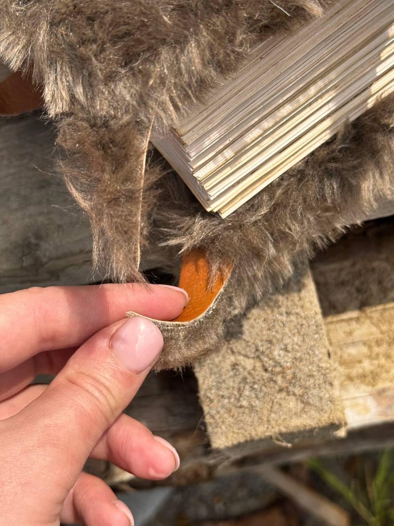 May include: Close-up of a section of brown faux fur with a stack of thin, light-colored wooden planks. The fur has a shaggy texture, and a small piece is being held up, revealing an orange underside.