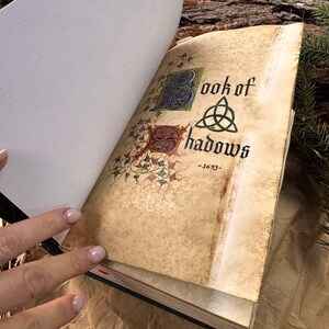 May include: An open book with a worn, aged cover. The title page reads "Book of Shadows - 1623" with a decorative triquetra symbol. The book is open to a blank page.