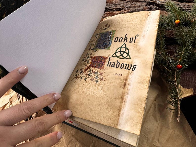 May include: An open book with a worn, aged cover. The title page reads "Book of Shadows - 1623" with a decorative triquetra symbol. The book is open to a blank page.