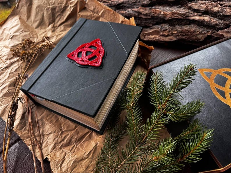 May include: Two black leather-bound books with a Celtic knot design on the cover. The book on the left has a red knot, and the book on the right has a gold knot. A pine branch is in the foreground.