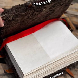 May include: A handmade, open book with a brown, furry cover, adorned with white teeth and brown leather accents. A red ribbon serves as a bookmark. The pages are blank and off-white, suggesting a journal or sketchbook.