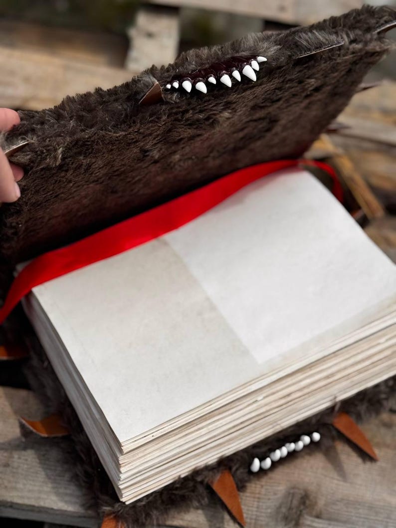May include: A handmade, open book with a brown, furry cover, adorned with white teeth and brown leather accents. A red ribbon serves as a bookmark. The pages are blank and off-white, suggesting a journal or sketchbook.