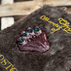 May include: A detailed close-up of a prop book with a furry, gray cover. A burgundy-colored, multi-eyed monster face is centered on the cover. The book has black and yellow text that reads "Monster of the Book" on a black patch.