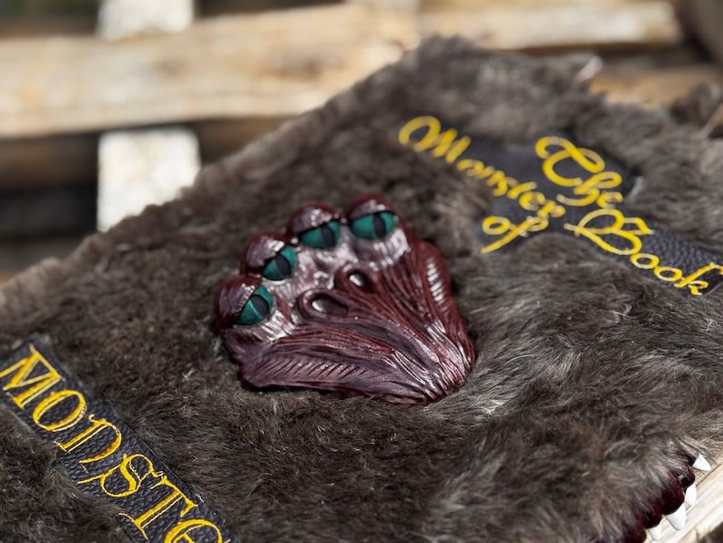May include: A detailed close-up of a prop book with a furry, gray cover. A burgundy-colored, multi-eyed monster face is centered on the cover. The book has black and yellow text that reads "Monster of the Book" on a black patch.
