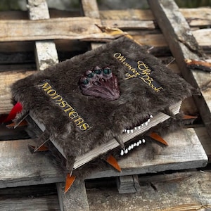 May include: A faux fur-covered book titled "The Monster Book of Monsters" with a monster claw clasp. The book has a toothy grin and orange spikes. The word "MONSTERS" is embroidered in gold.